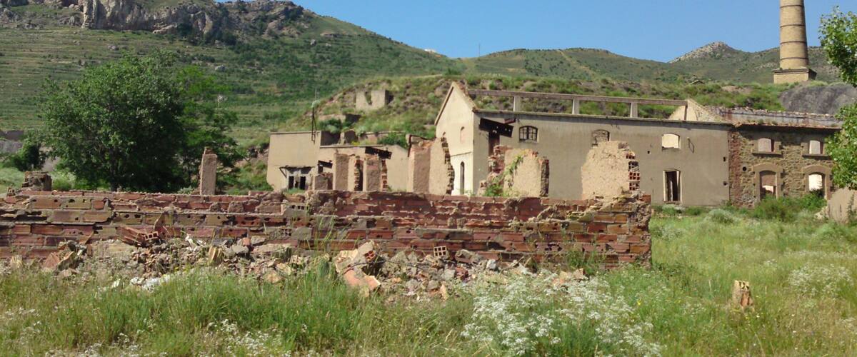 A dilapidated industrial site with a chimney, scattered ruins, and a mountainous backdrop.
