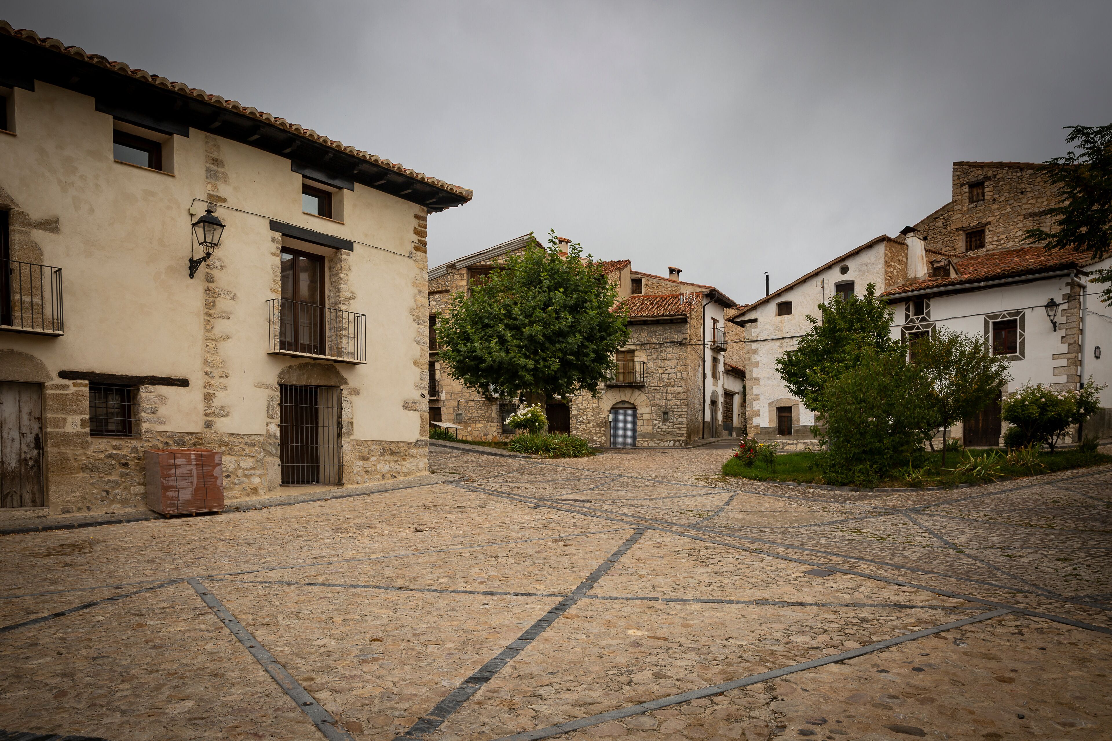 A cobblestone street in a historic town with stone buildings, a large tree, and a traditional lamp post.