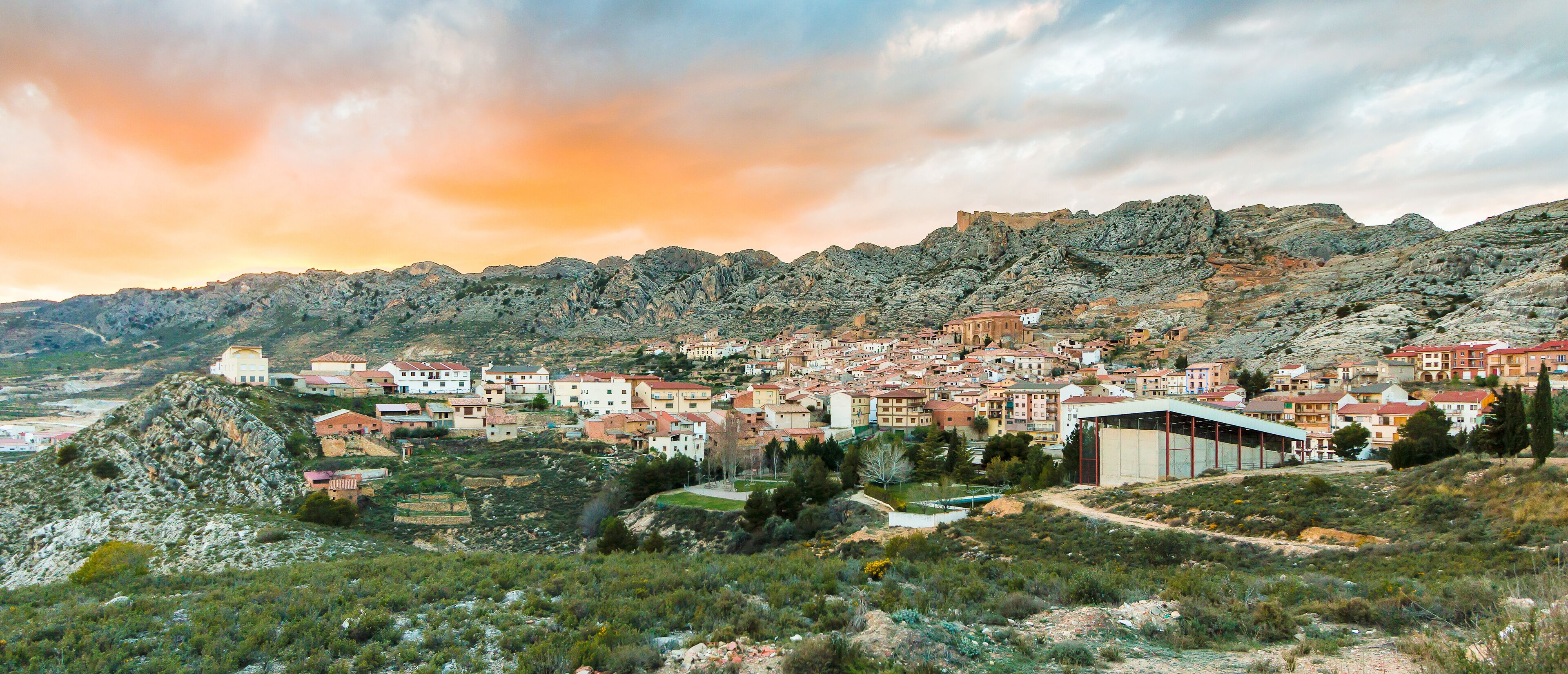 A coastal town with red-roofed buildings, a rocky hillside, and a clear sky.