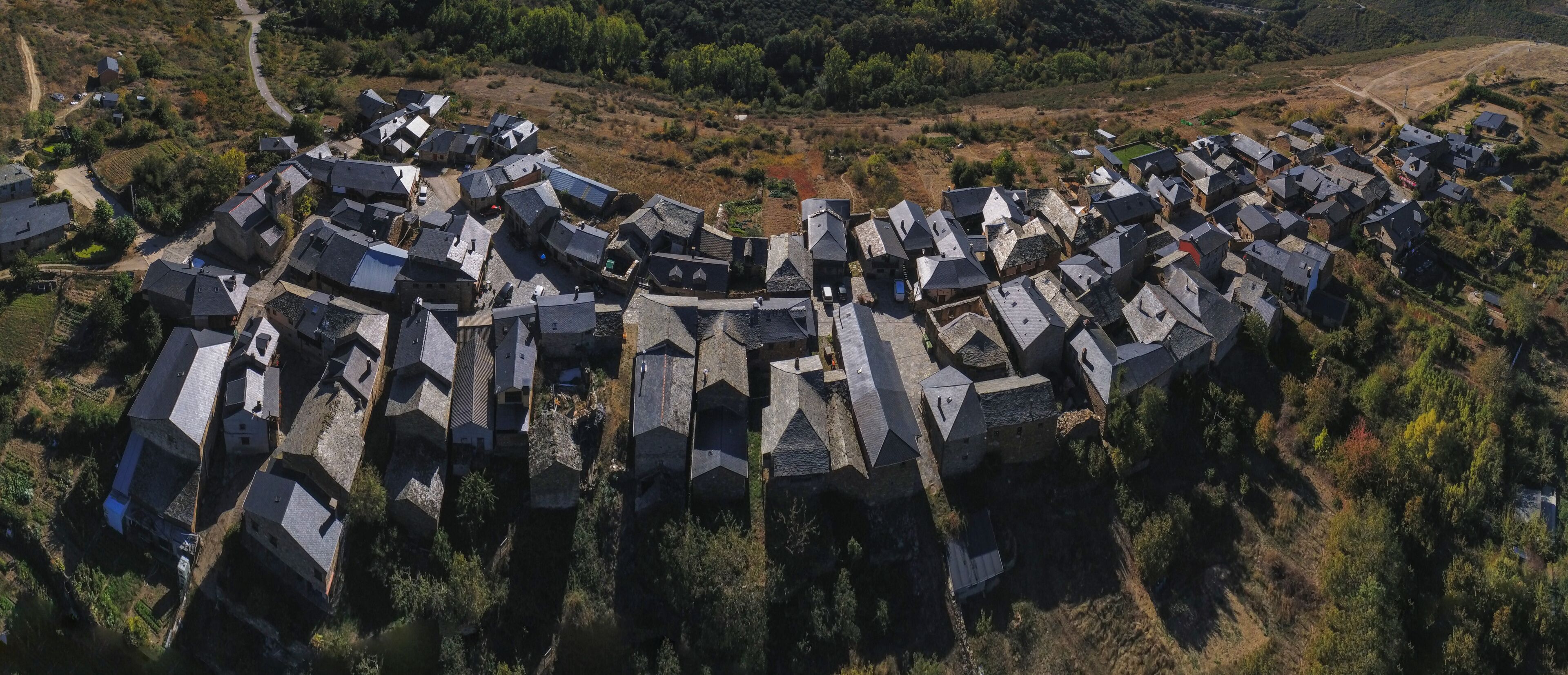 An aerial view of a village with stone houses arranged in a circular pattern, surrounded by forested hills.
