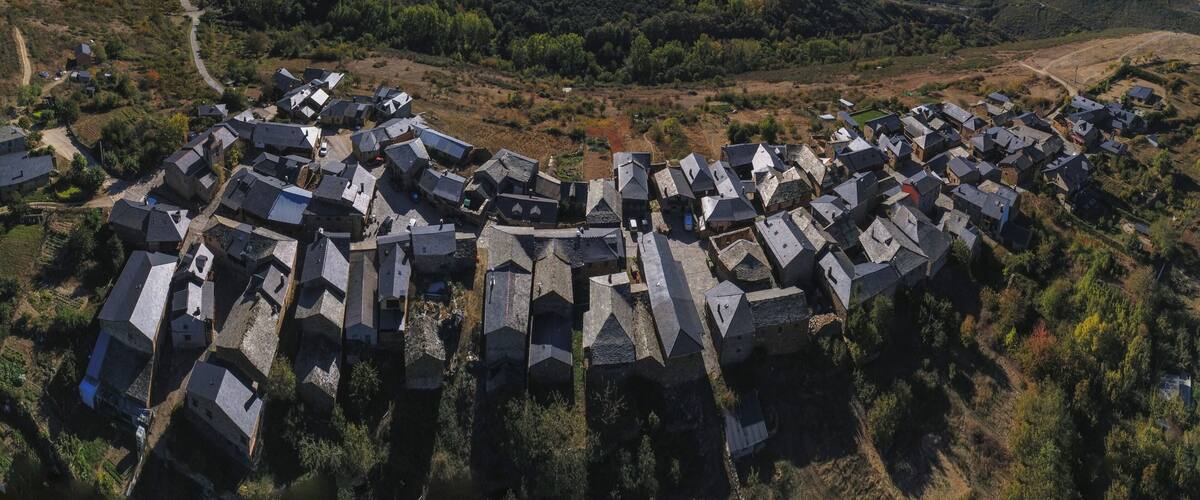An aerial view of a village with stone houses arranged in a circular pattern, surrounded by forested hills.