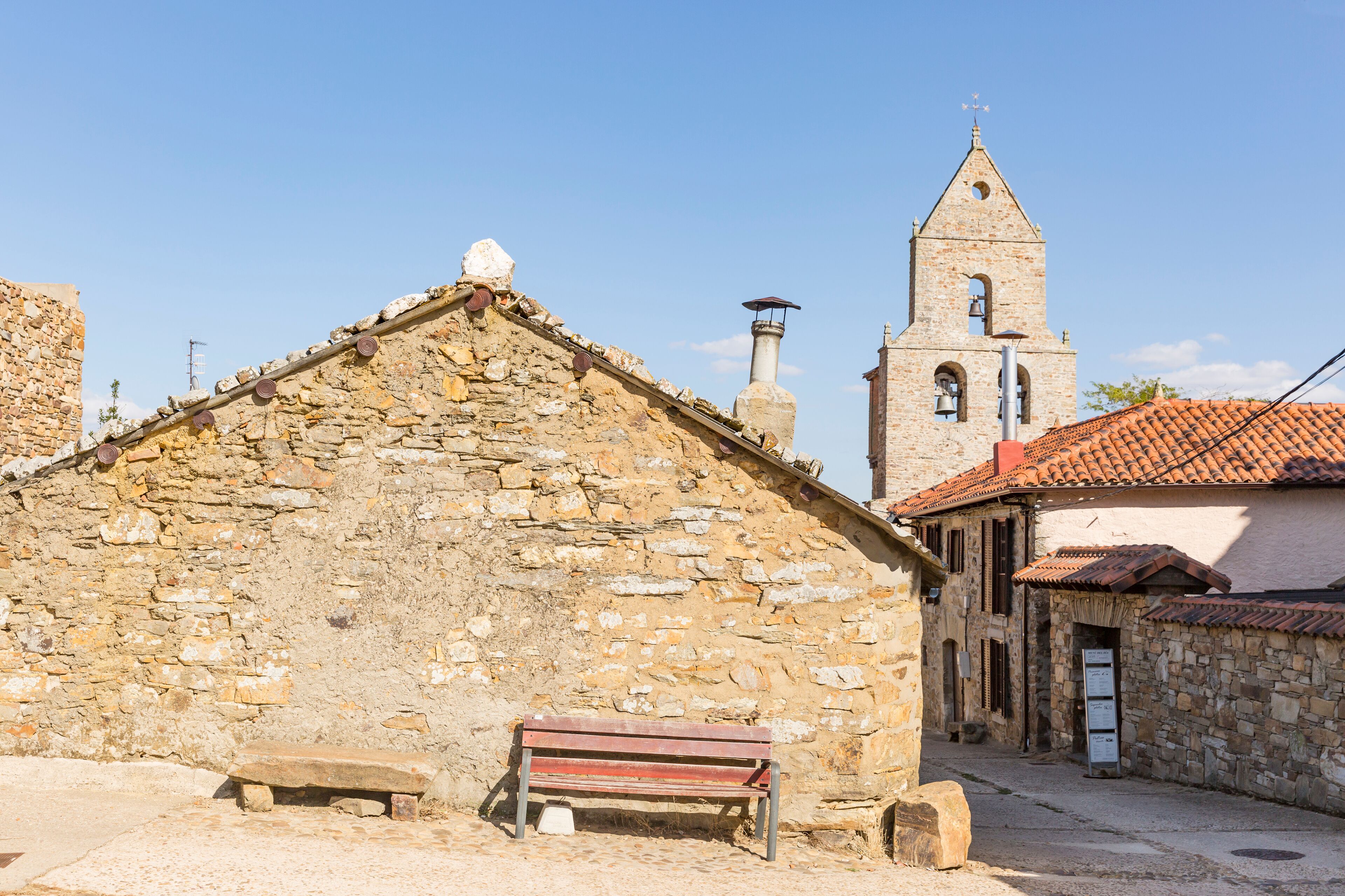 A stone building with a red bench in front, a church with a bell tower, and a clear blue sky.