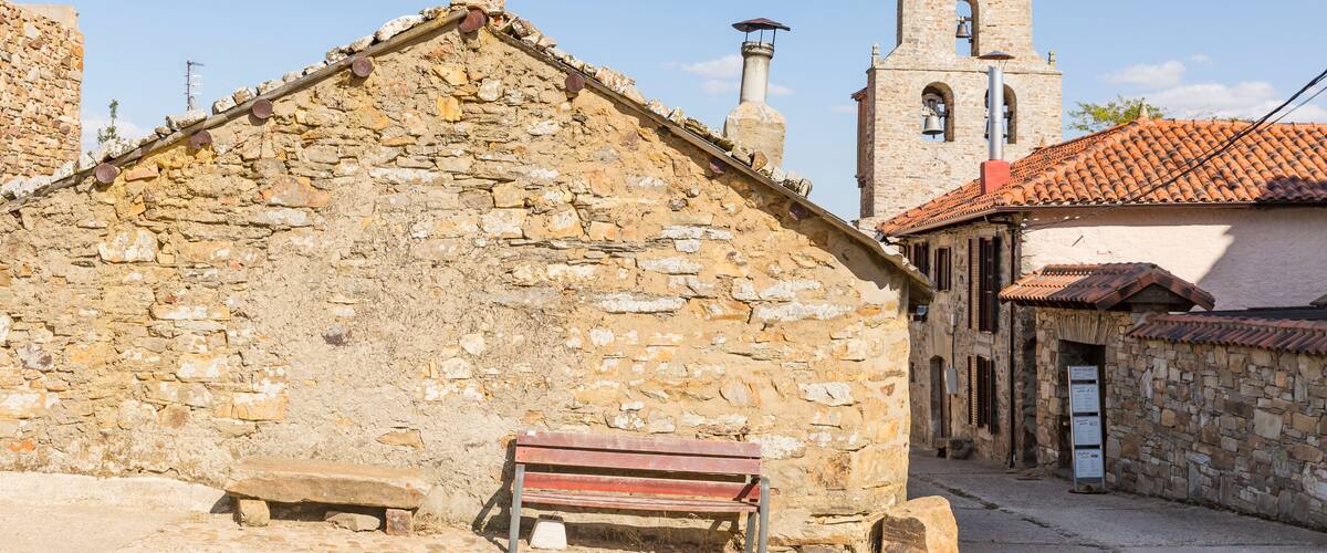 A stone building with a red bench in front, a church with a bell tower, and a clear blue sky.