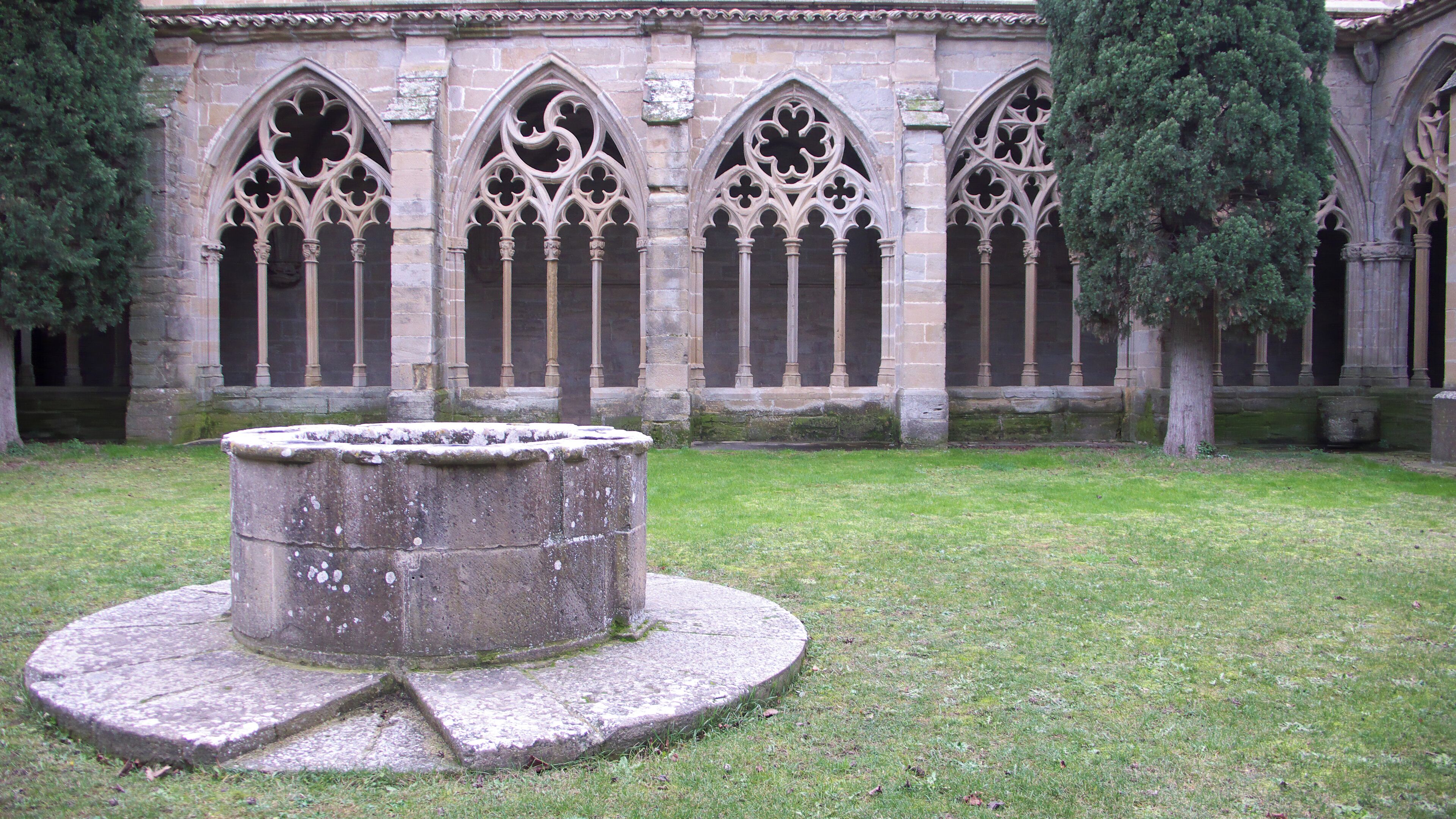 A medieval cloister with a central fountain, stone arches, and a grassy courtyard.