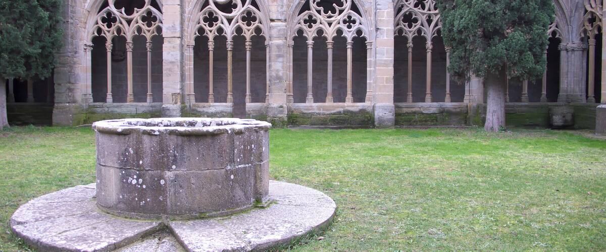 A medieval cloister with a central fountain, stone arches, and a grassy courtyard.