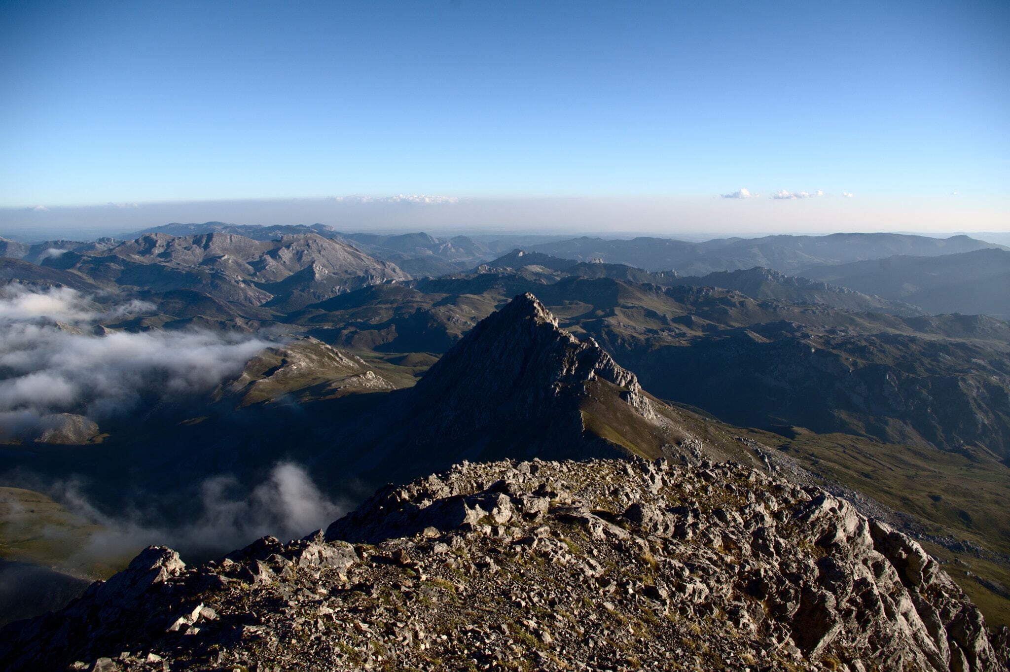 Un pico montañoso con terreno rocoso y cordilleras lejanas bajo un cielo despejado.