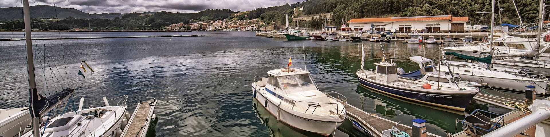 A marina with boats docked, a wooden pier, and a backdrop of a hilly landscape with buildings.