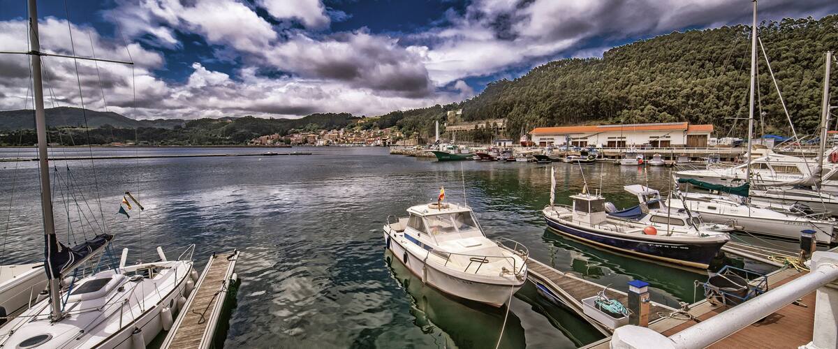 A marina with boats docked, a wooden pier, and a backdrop of a hilly landscape with buildings.