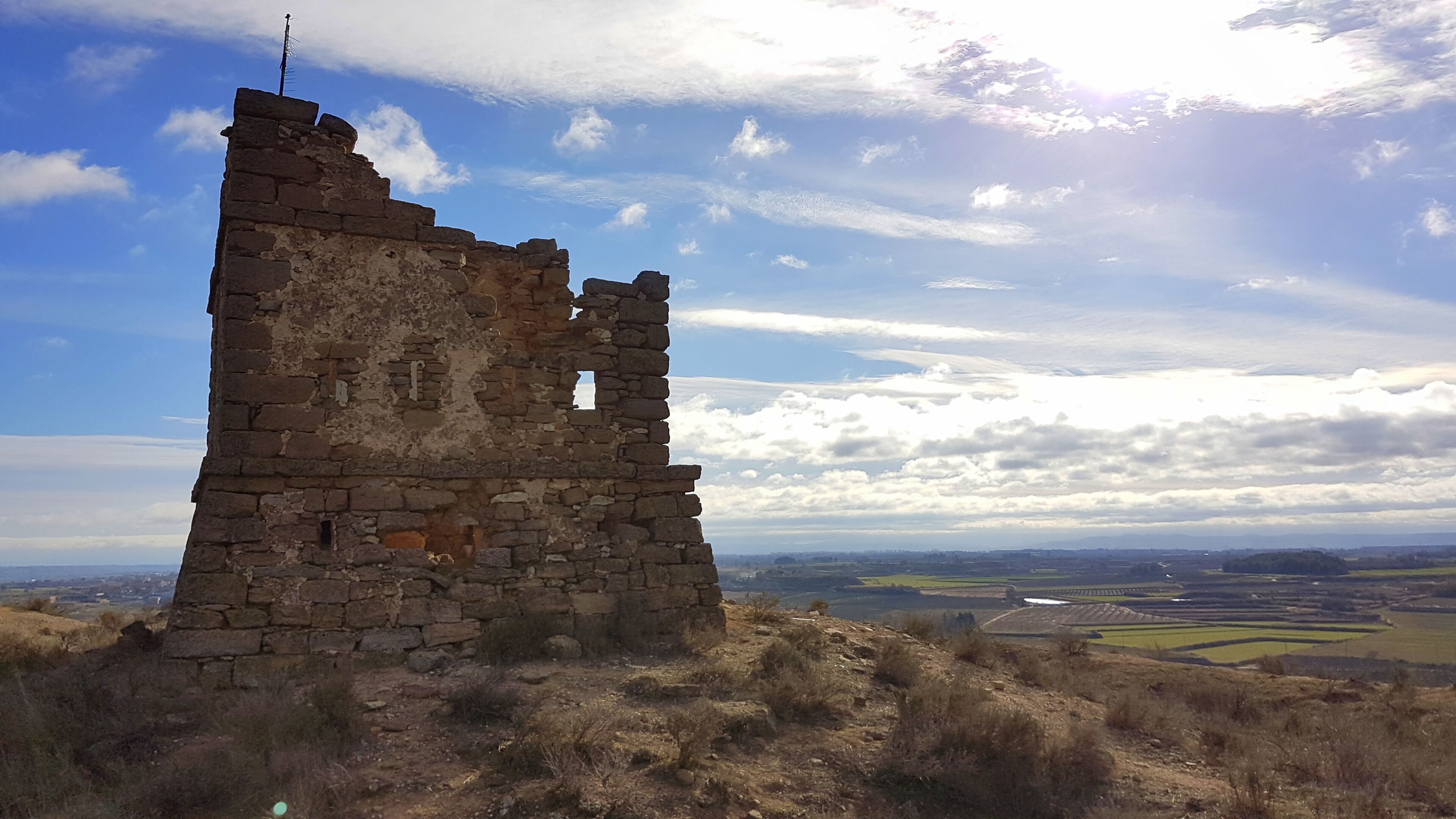 A stone ruin with a cross on top, surrounded by a hilly landscape and a partly cloudy sky.