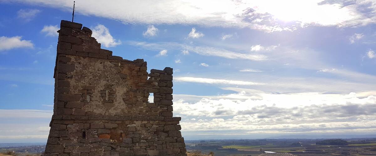 A stone ruin with a cross on top, surrounded by a hilly landscape and a partly cloudy sky.