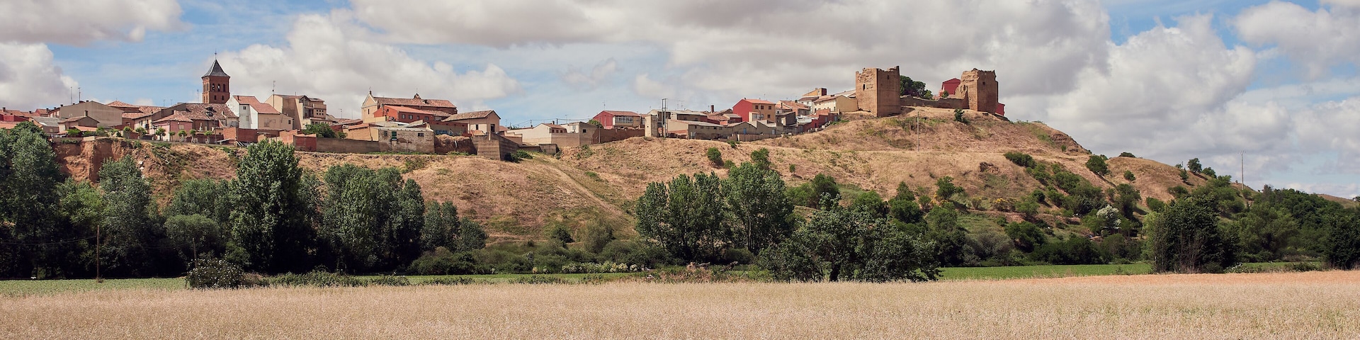 A historic town with medieval architecture, including a prominent tower and castle ruins, set against a backdrop of rolling hills and a clear sky.