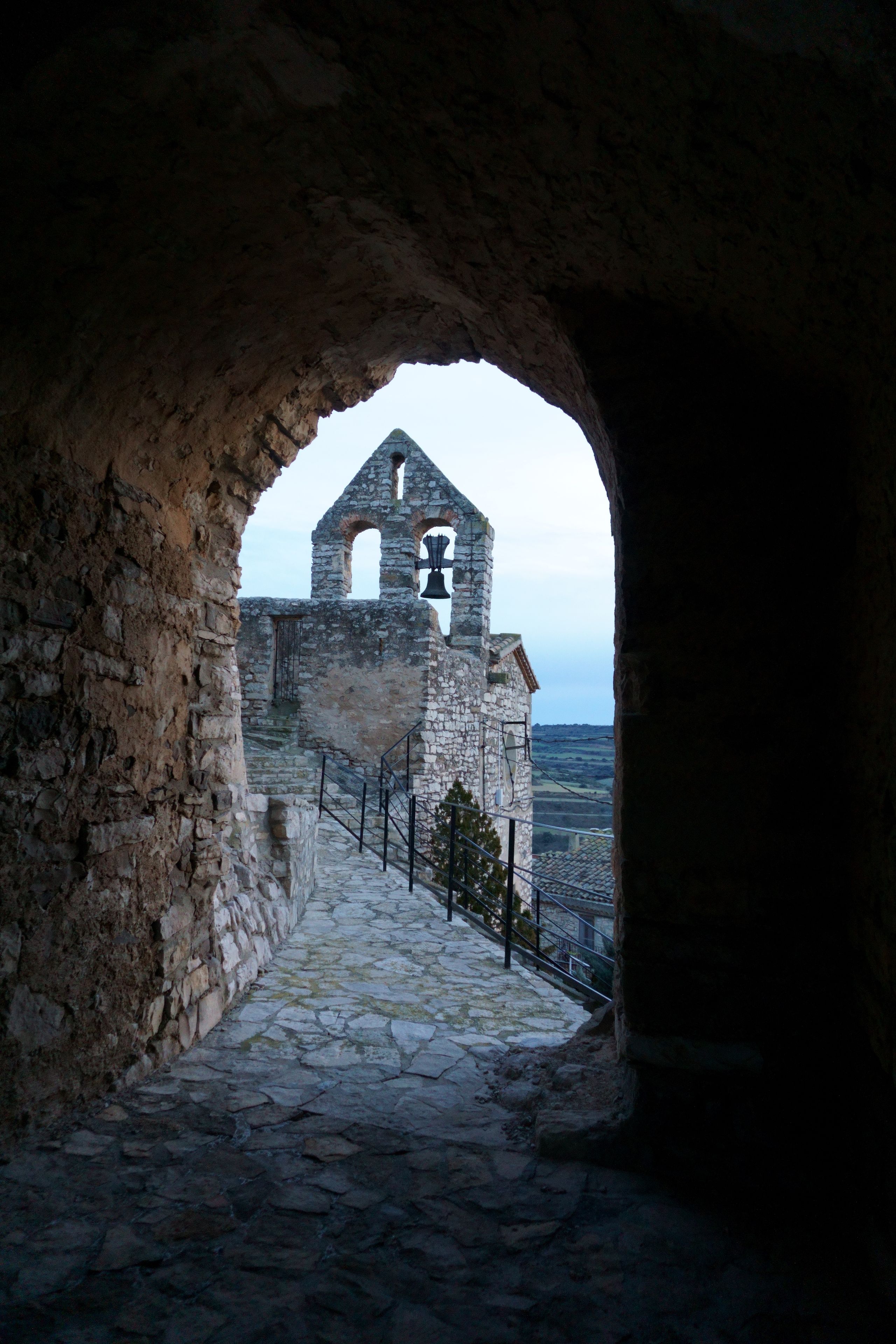 A stone pathway leading to a historic stone building with a bell tower, viewed through an arched opening.