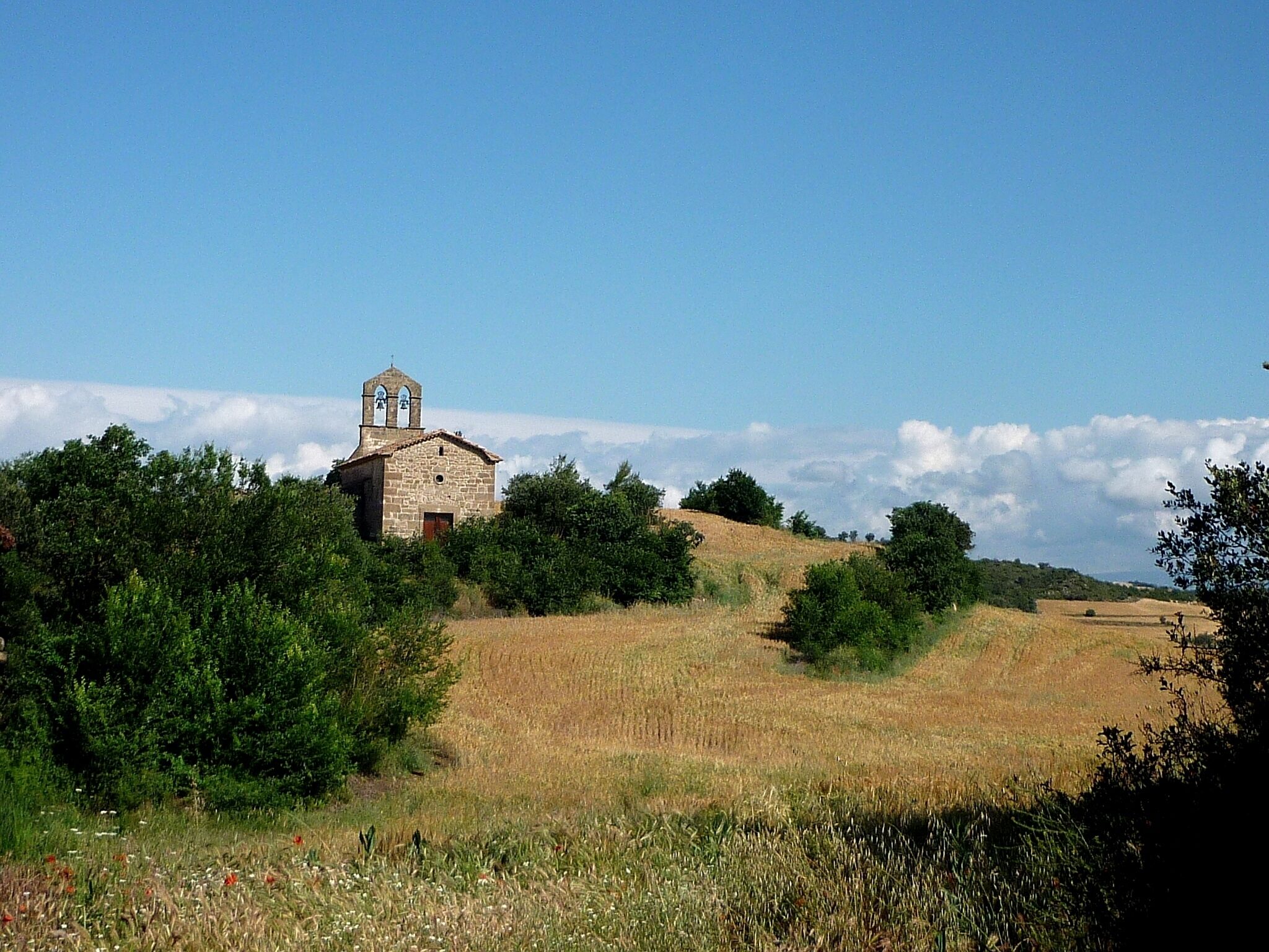 A stone church with a bell tower on a hill, surrounded by fields and trees.