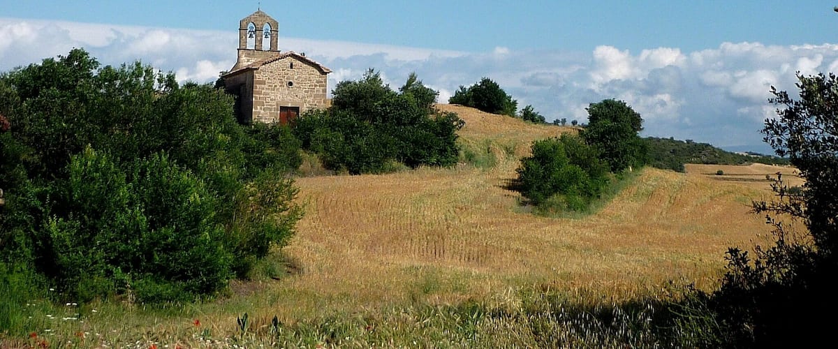 A stone church with a bell tower on a hill, surrounded by fields and trees.