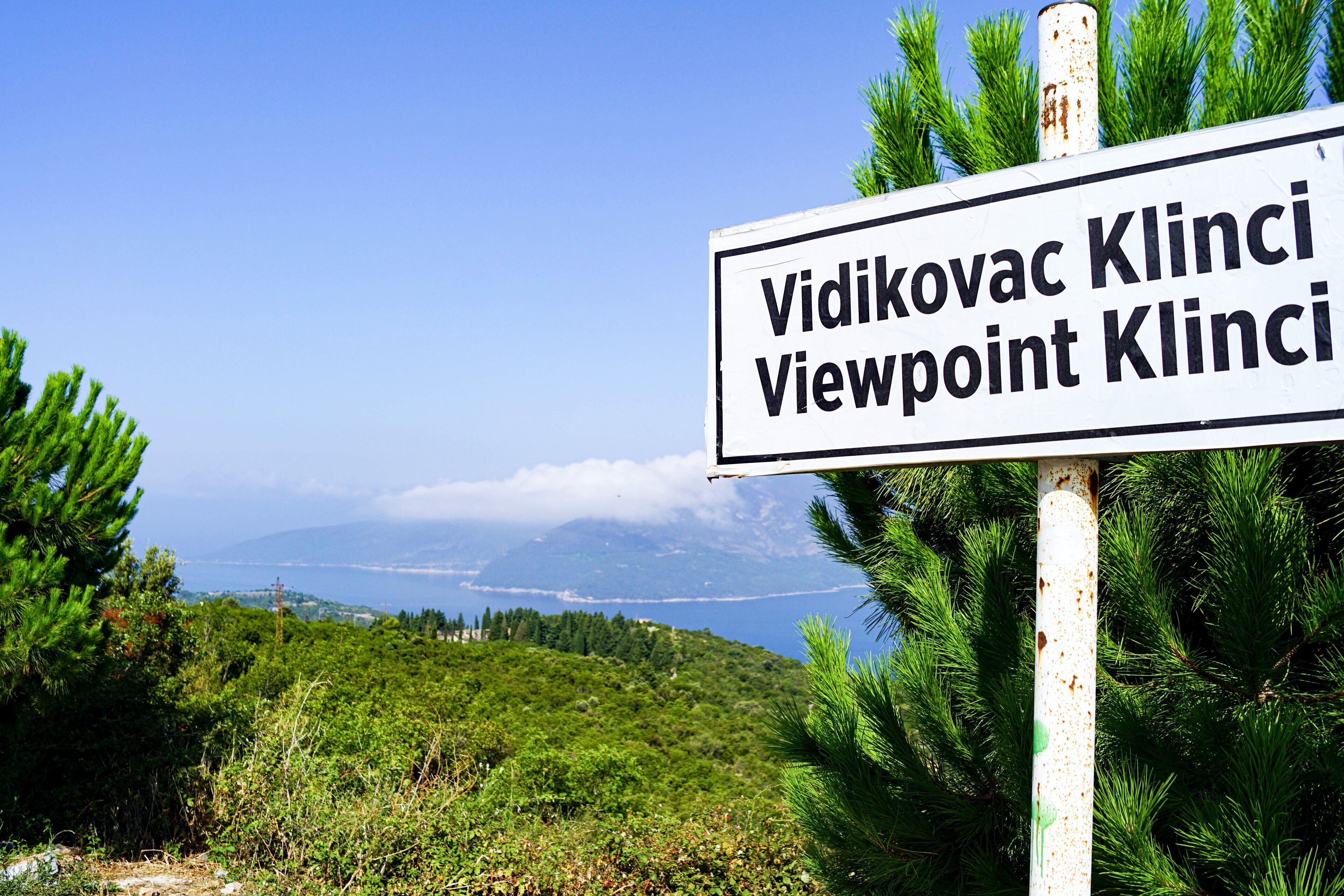 A sign with 'Vidikovac Klinci Viewpoint Klinci' text, surrounded by greenery and a scenic view of a coastline.