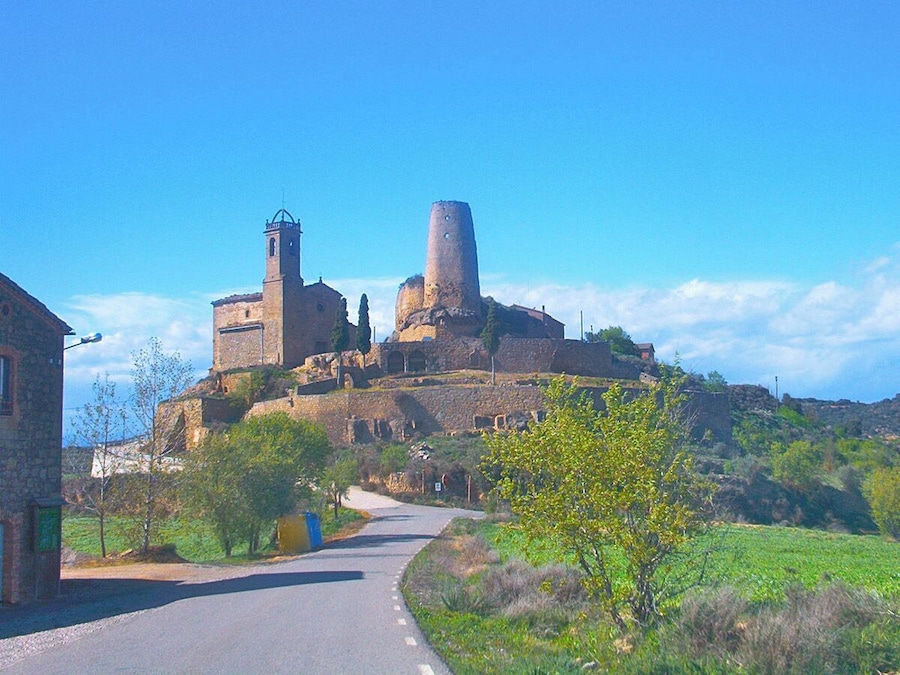 Una fortalesa de pedra històrica, amb una torre campanar i una torre cilíndrica, envoltada de vegetació i una carretera.