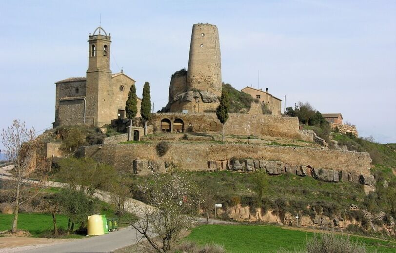 Uma fortaleza de pedra histórica com uma torre de sino e uma torre cilíndrica, rodeada por vegetação e uma estrada pavimentada.