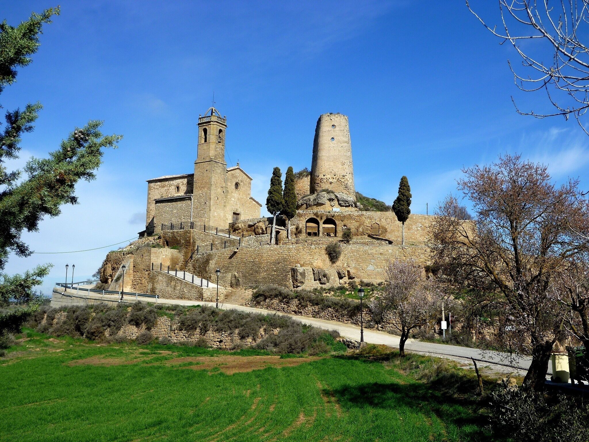 A historic stone building with a bell tower and a tall cylindrical structure, surrounded by greenery and a paved path.