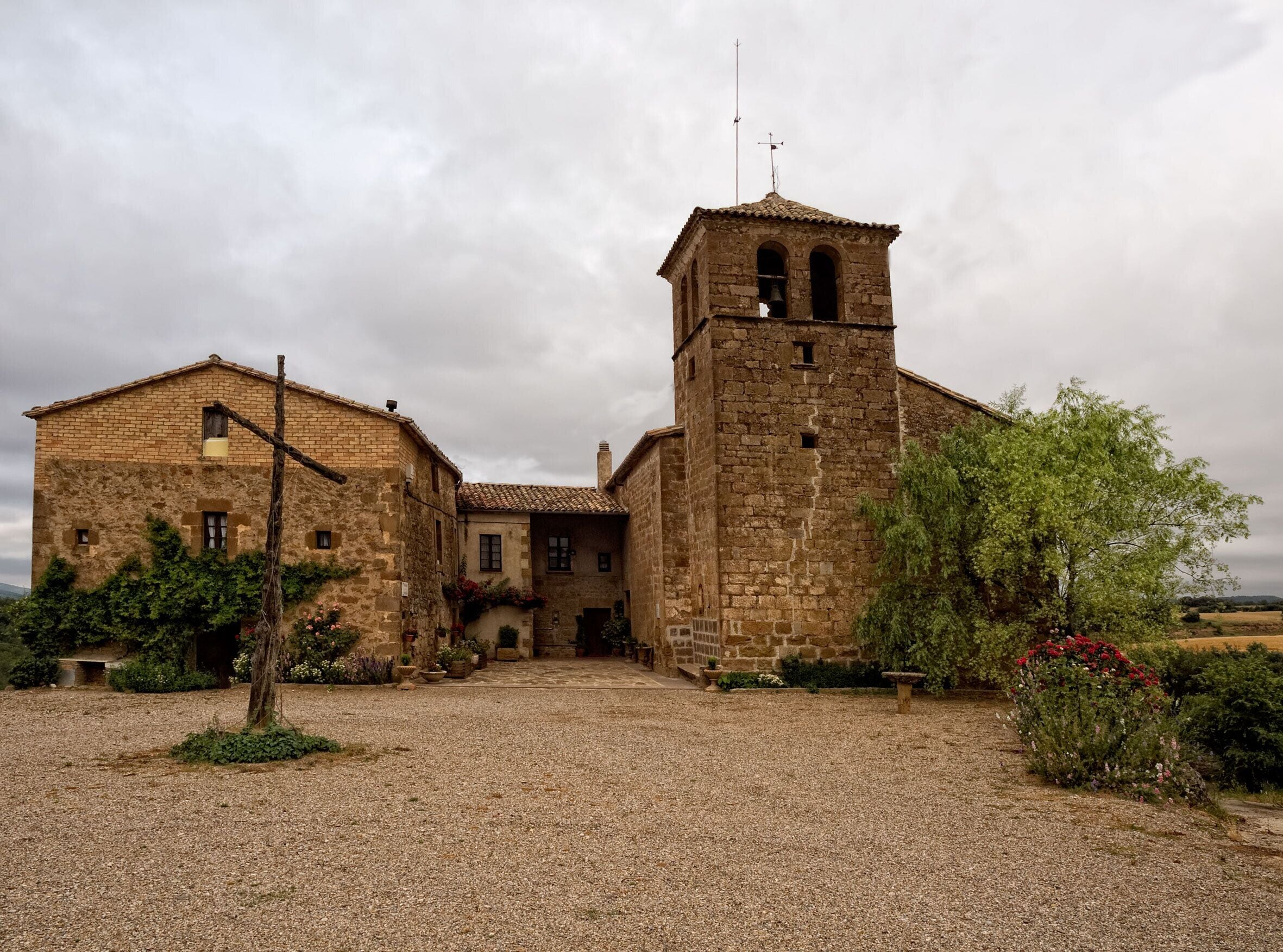 A historic stone building with a bell tower, surrounded by gravel and greenery.