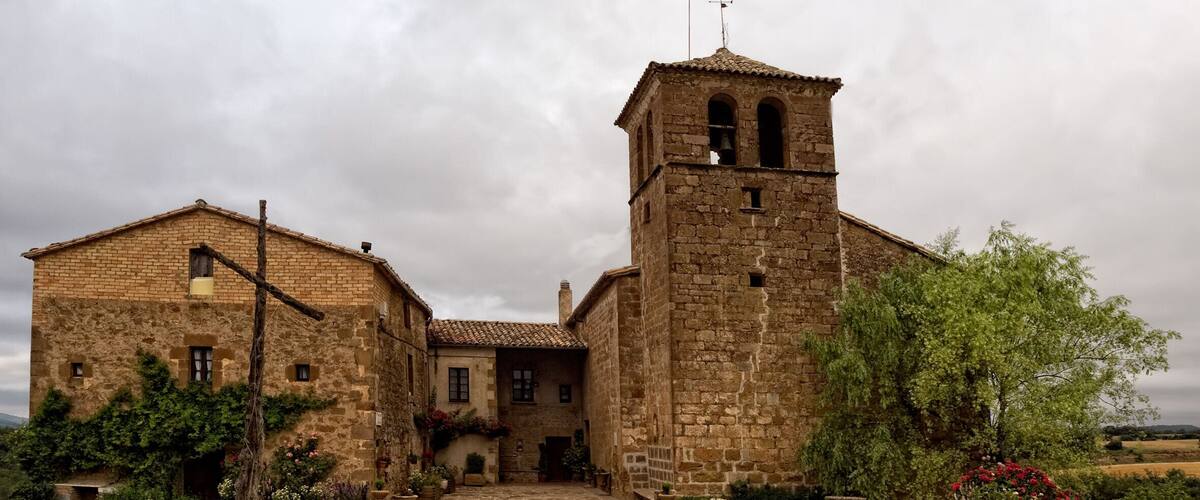 A historic stone building with a bell tower, surrounded by gravel and greenery.
