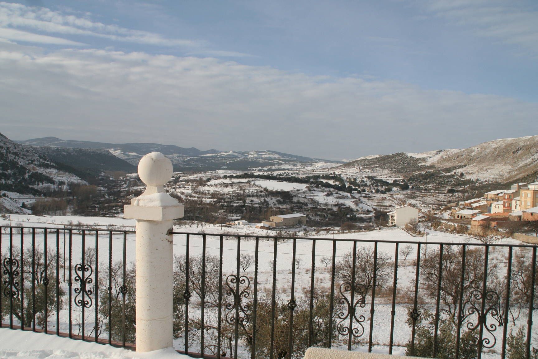 A snow-covered landscape with a mountainous backdrop, a fenced area with a decorative post, and a small settlement with buildings.