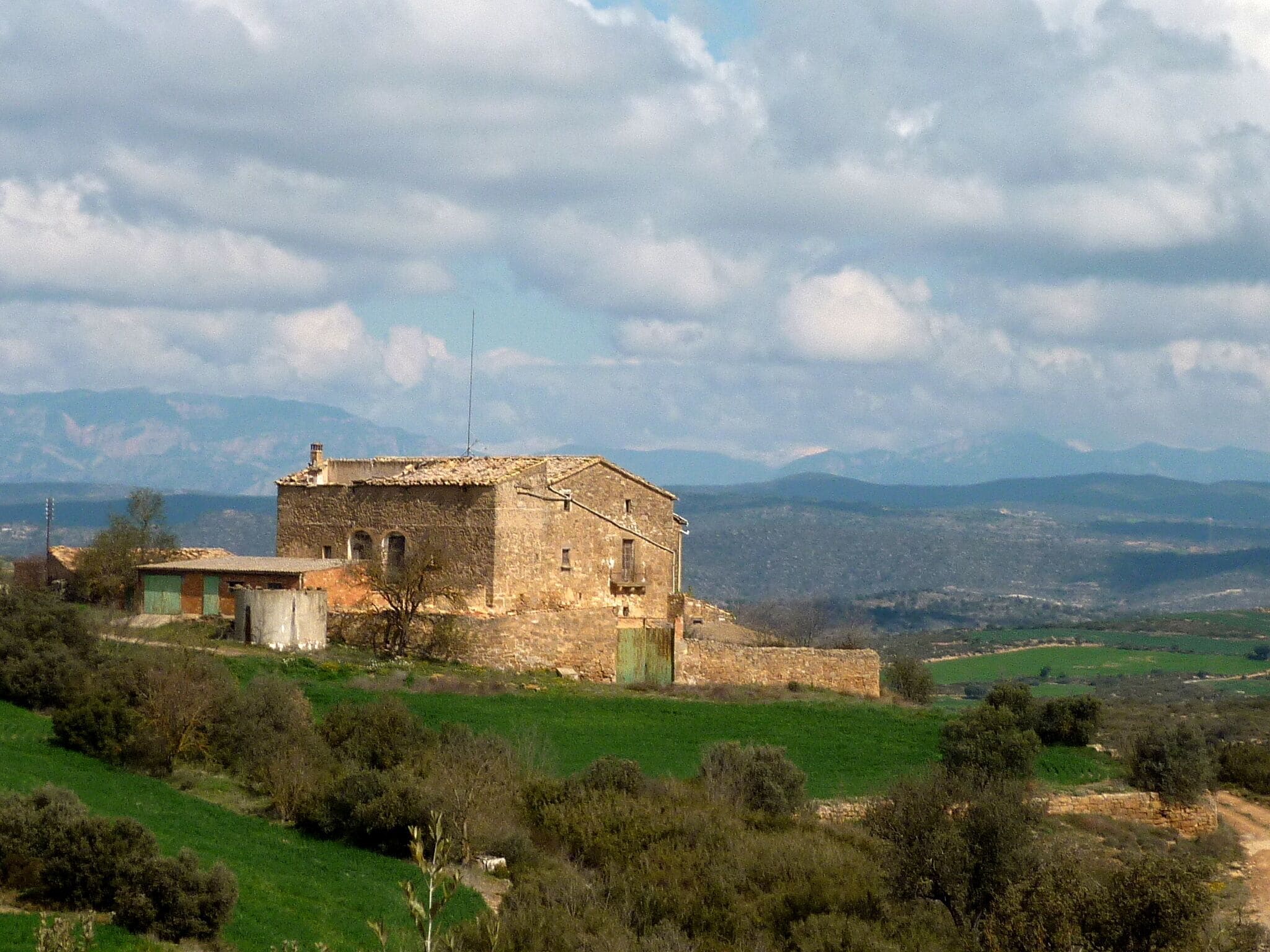 A stone building with a tiled roof surrounded by green fields and mountains in the background.