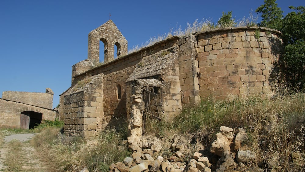 An ancient stone building with arched windows and a bell tower, surrounded by dry grass and rocks.