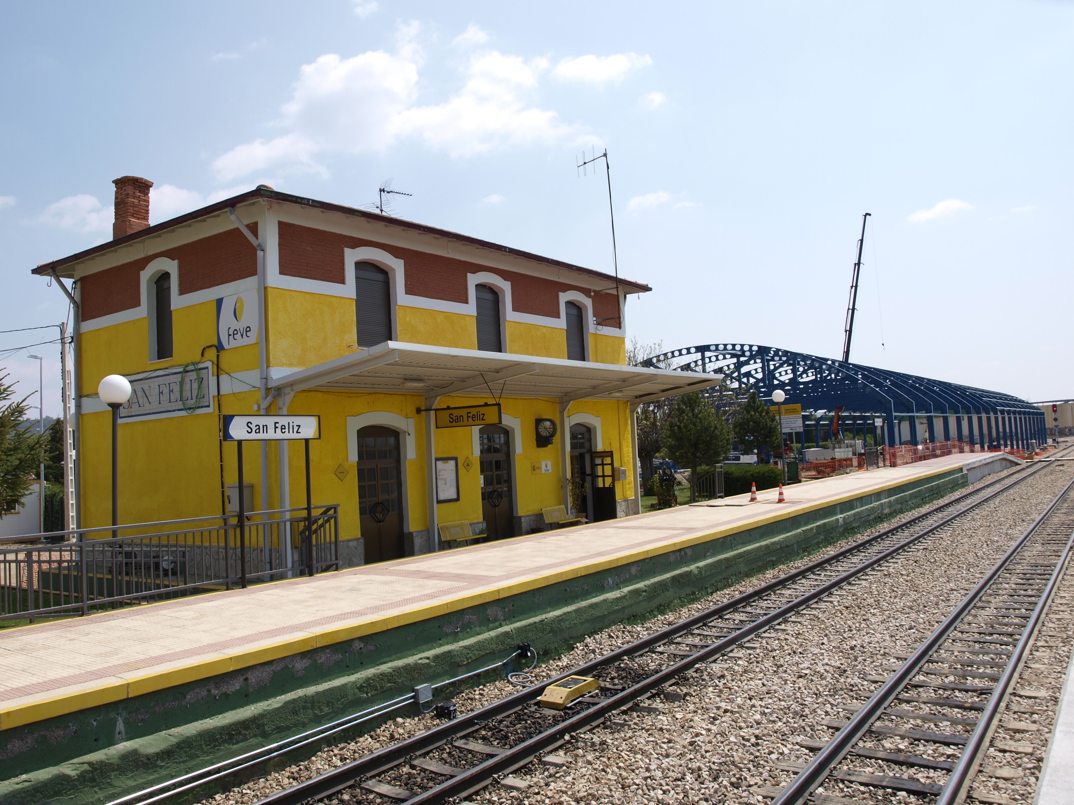 A train station named San Feliz with a yellow building, a covered platform, and a roller coaster in the background.