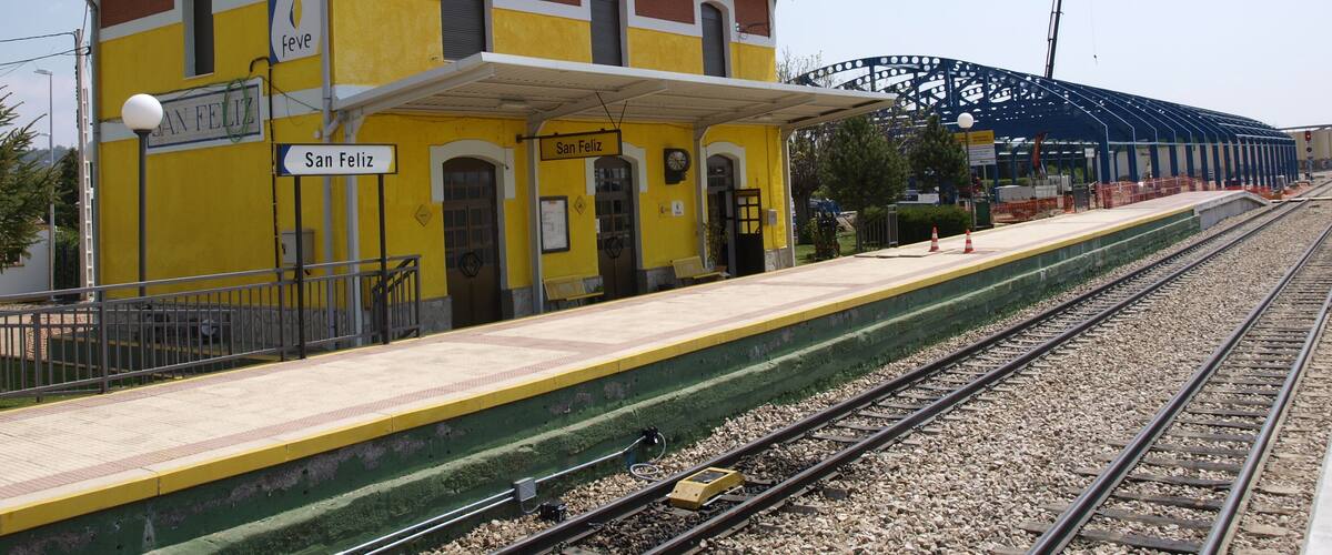 A train station named San Feliz with a yellow building, a covered platform, and a roller coaster in the background.