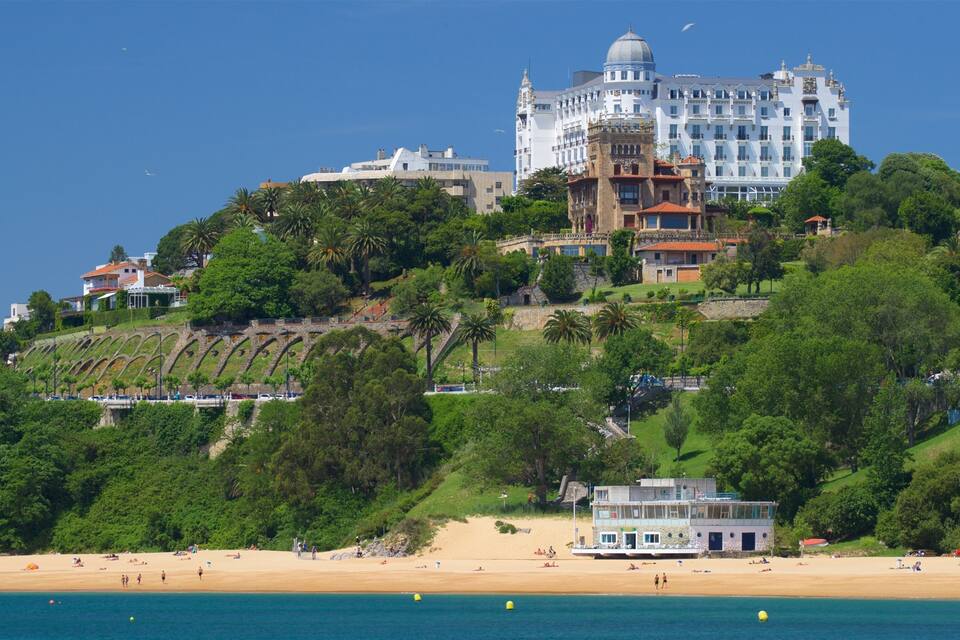 A coastal town with a prominent white building with a dome, lush green hills, and a sandy beach with people.