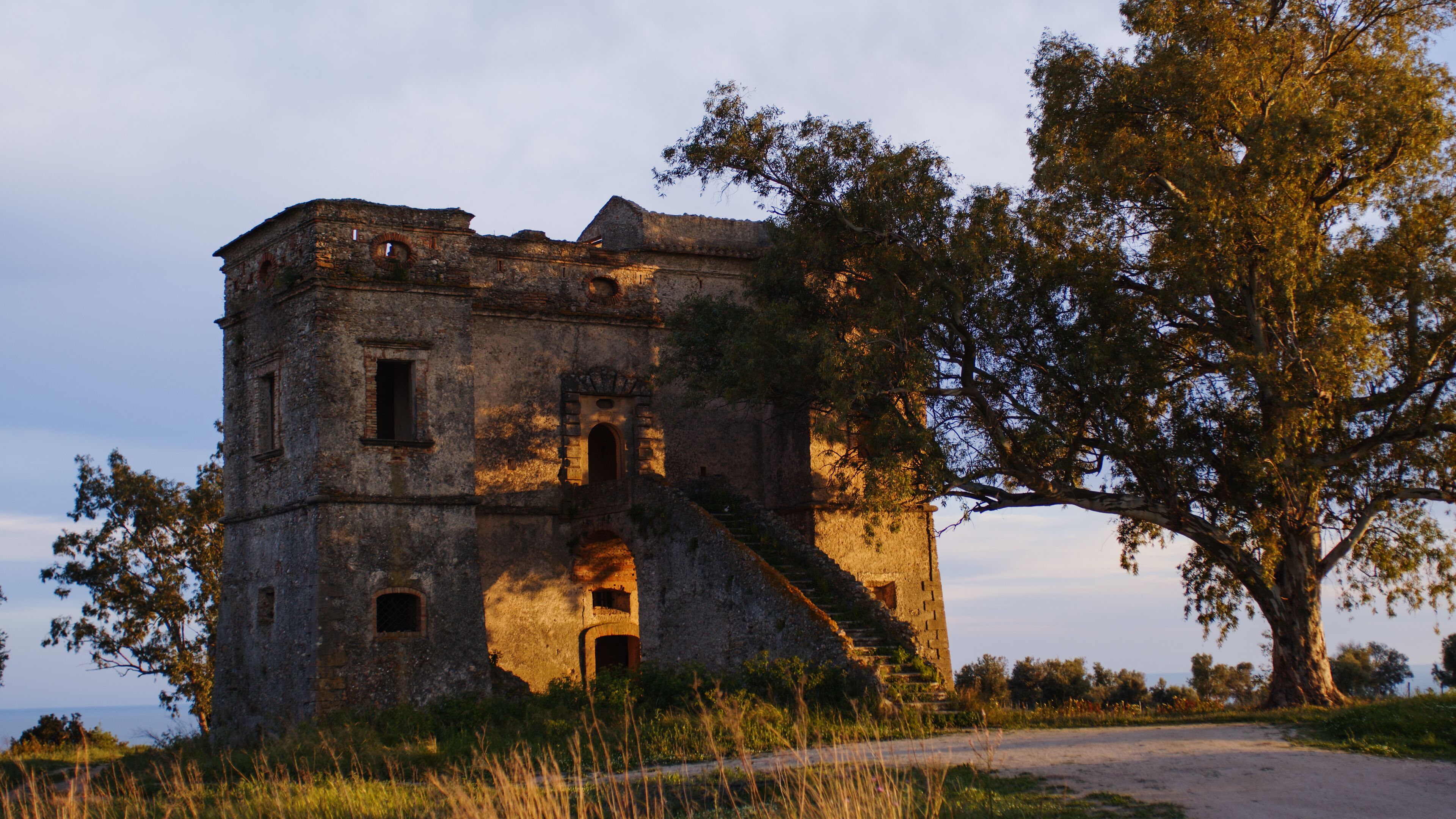 An old stone building with a staircase leading up to a door, surrounded by trees and grass.