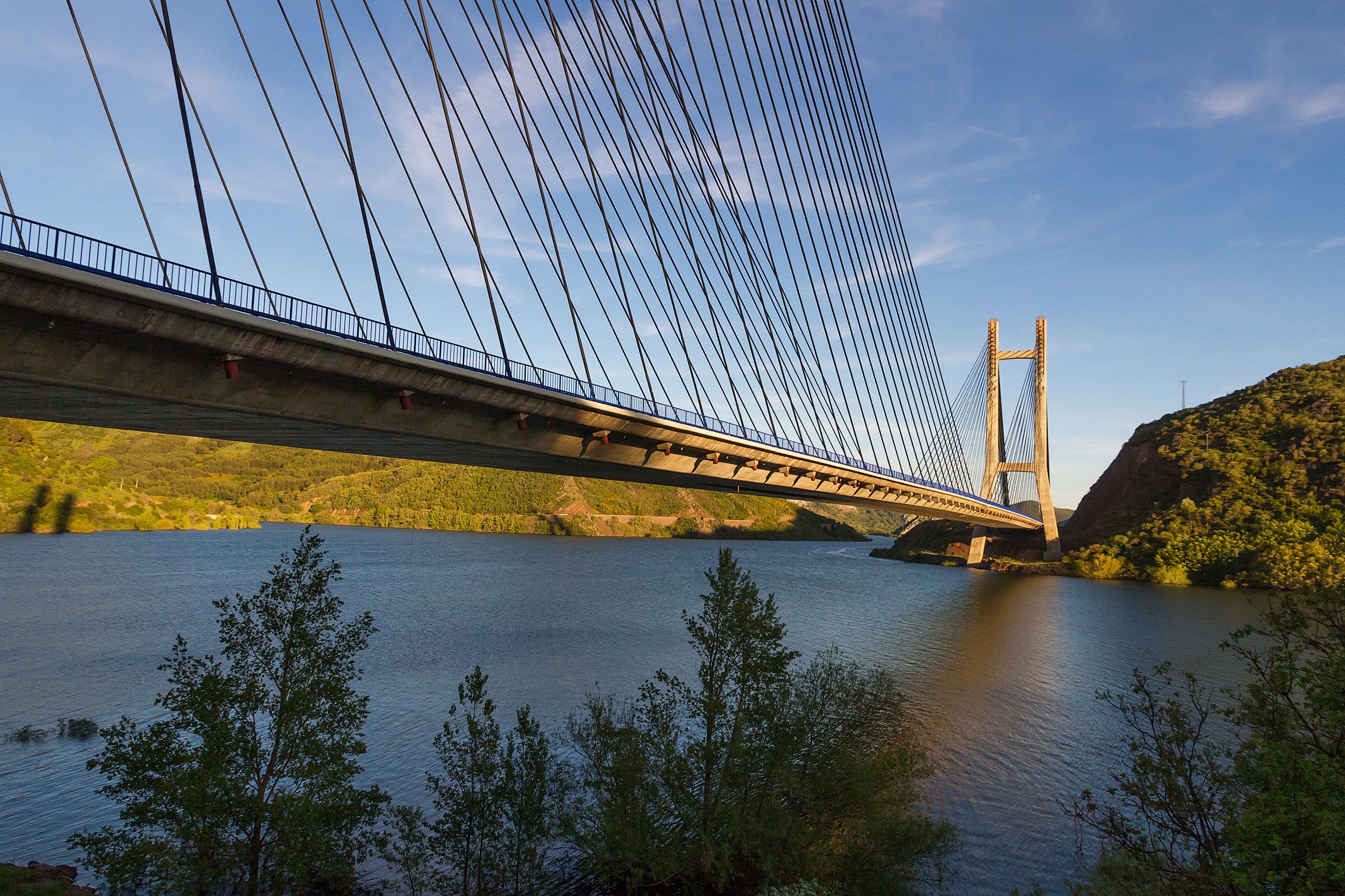 A suspension bridge with cables and towers over a river, surrounded by greenery and hills.