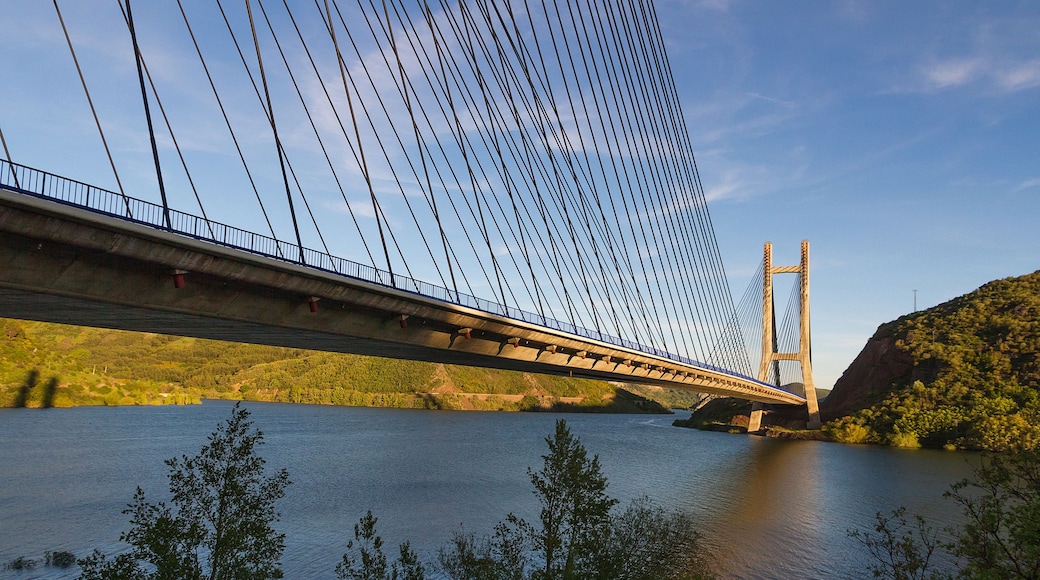 A suspension bridge with cables and towers over a river, surrounded by greenery and hills.