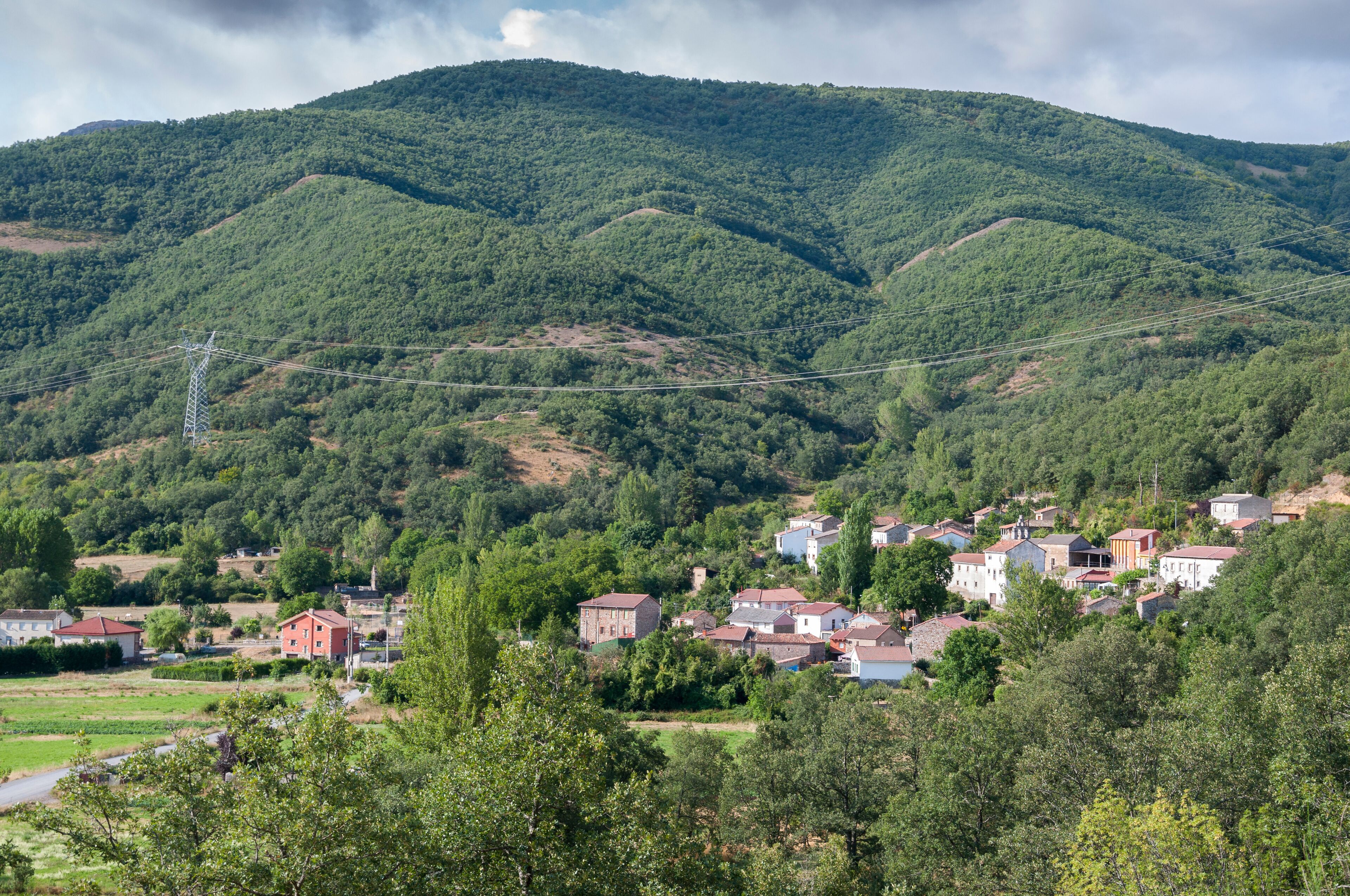 A village nestled in a valley with a backdrop of lush green hills and a power line running across the landscape.