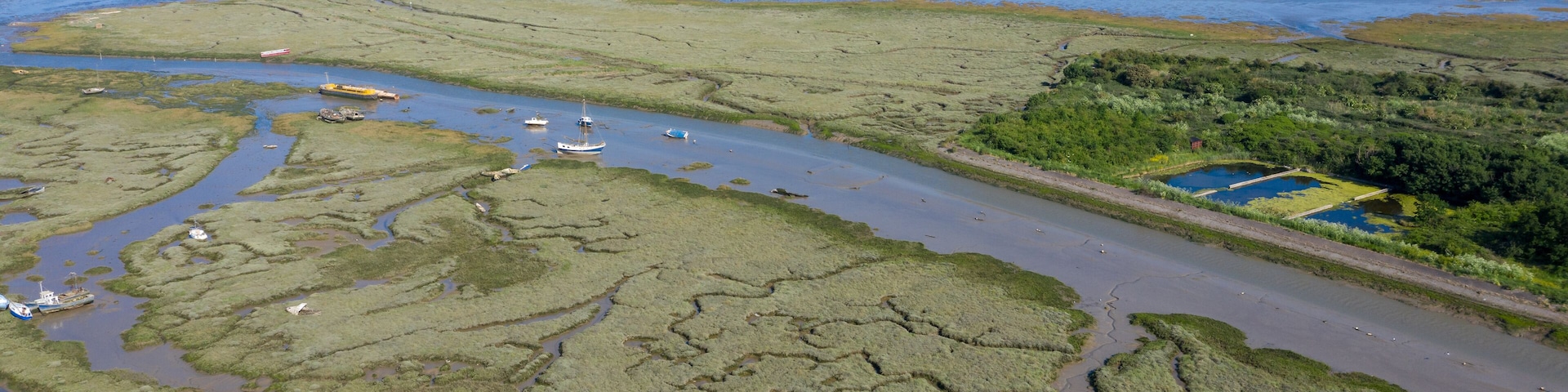 Paisagem costeira com um rio serpenteando por áreas pantanosas, barcos na água e o horizonte de uma cidade ao longe.
