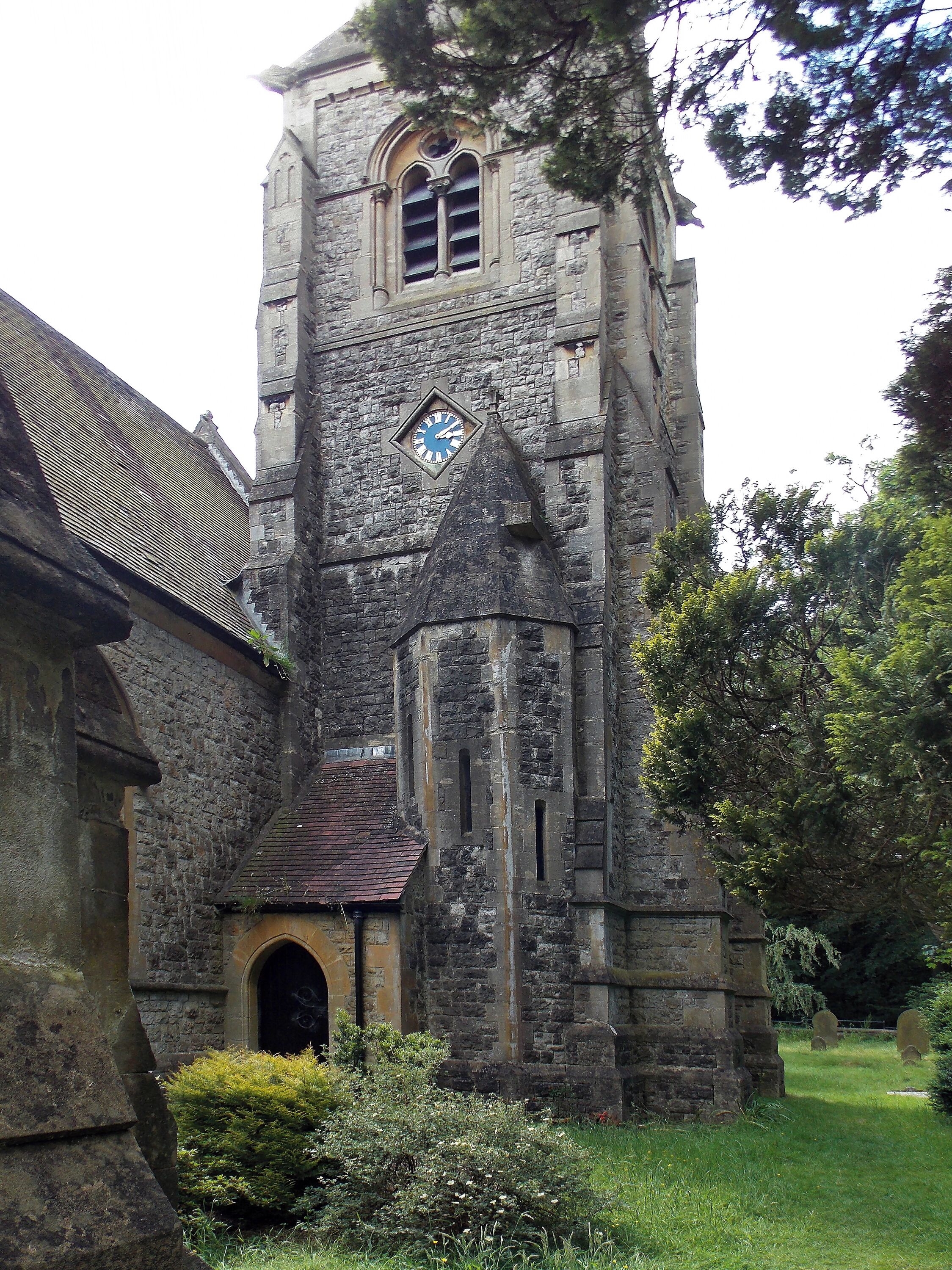 A historic stone church with a clock tower, surrounded by greenery and a cemetery.
