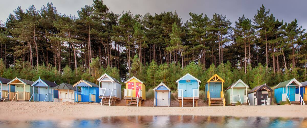 A row of colorful beach huts along a sandy beach with a forest in the background.