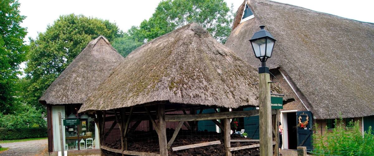 A traditional thatched-roof structure with a wooden frame, a brick building with a green sign, and a cobblestone path.