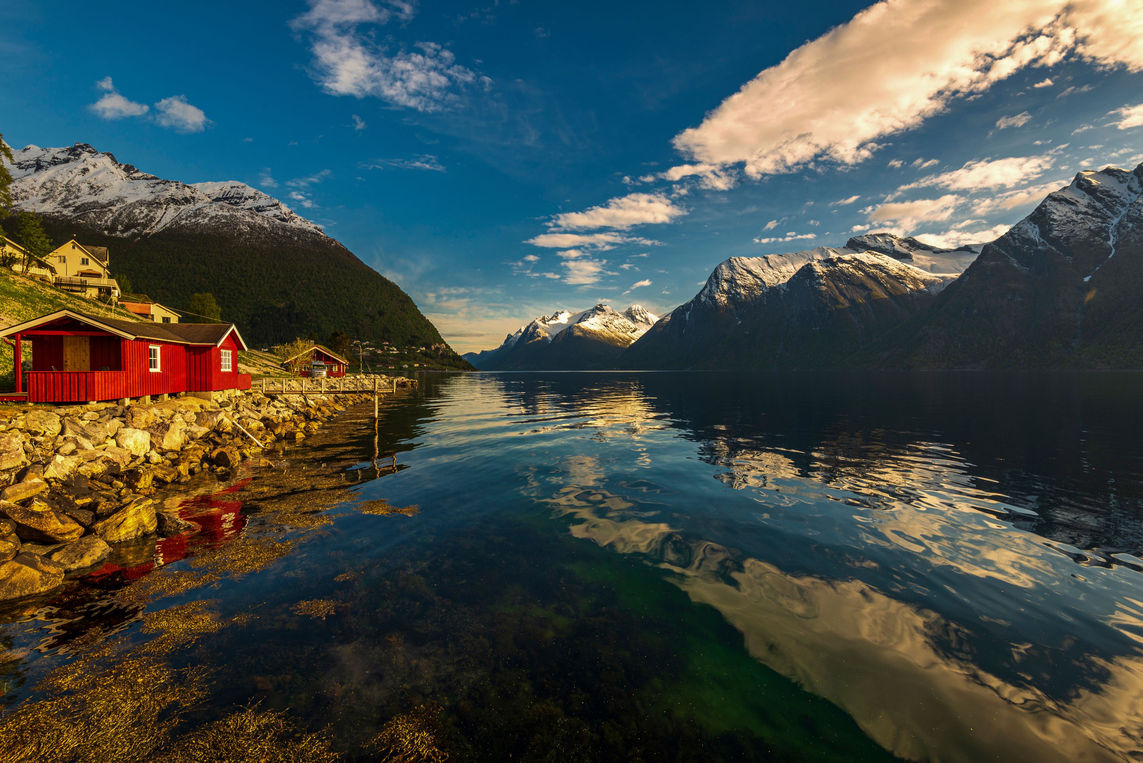 A calm fjord with a red wooden house on the left, snow-capped mountains in the background, and a clear blue sky.