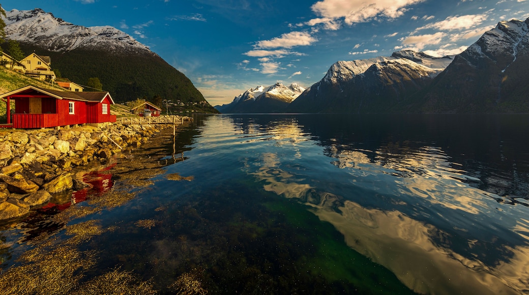 A calm fjord with a red wooden house on the left, snow-capped mountains in the background, and a clear blue sky.