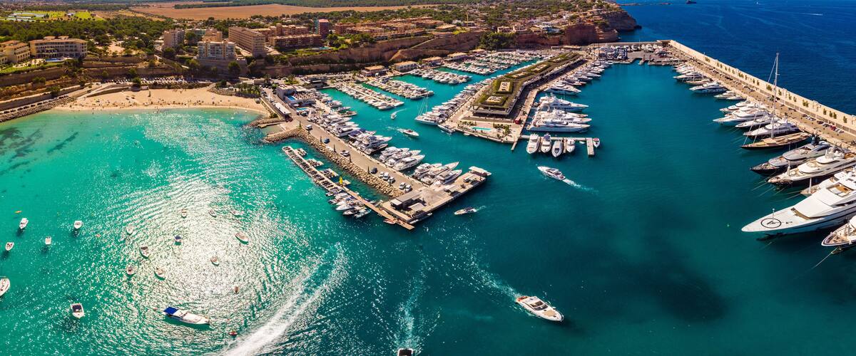 A coastal marina with numerous boats docked, a sandy beach, and a backdrop of hills and a clear blue sky.