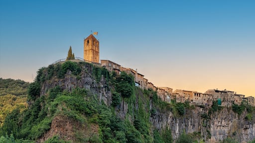 Uma torre de pedra histórica no topo de um penhasco, rodeada por vegetação exuberante e edifícios de pedra tradicionais.