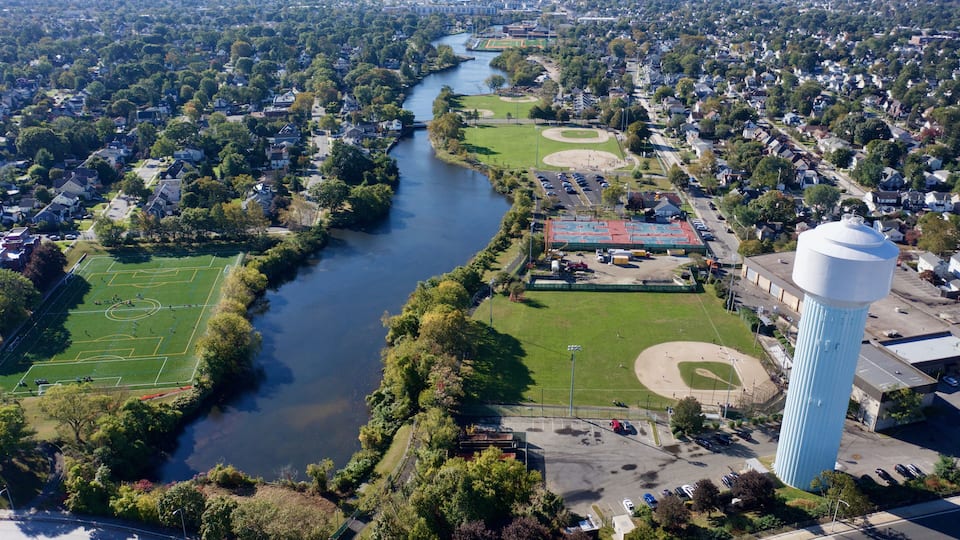 An aerial view of a river running through a residential area with sports fields and a water tower.