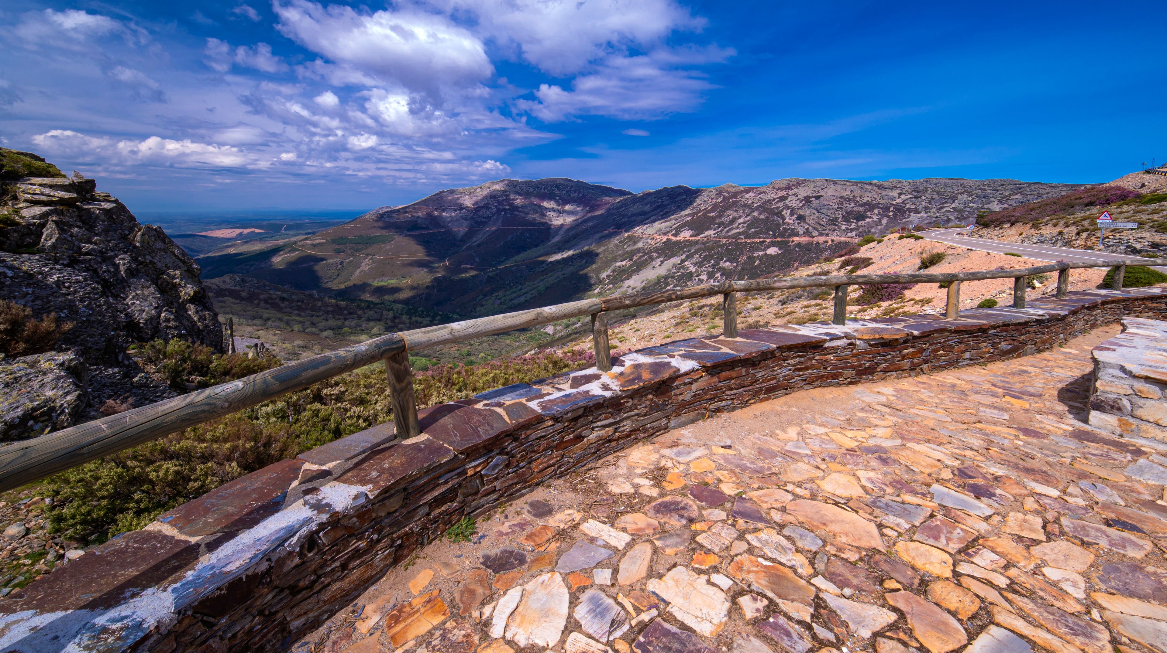 A stone pathway with a wooden railing, leading to a mountainous landscape with a clear sky.