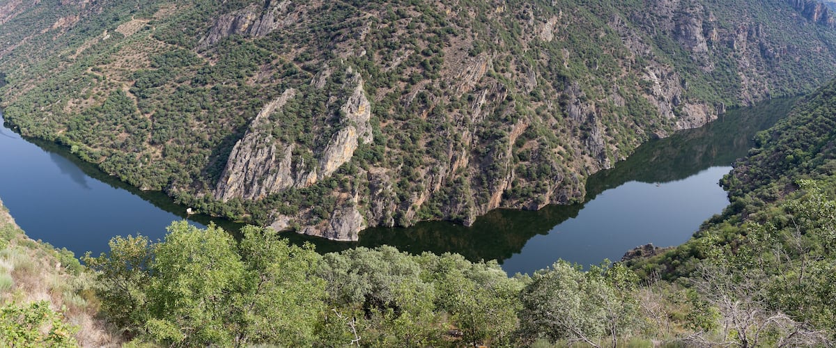 A mountainous landscape with a lake surrounded by dense greenery and rocky cliffs.