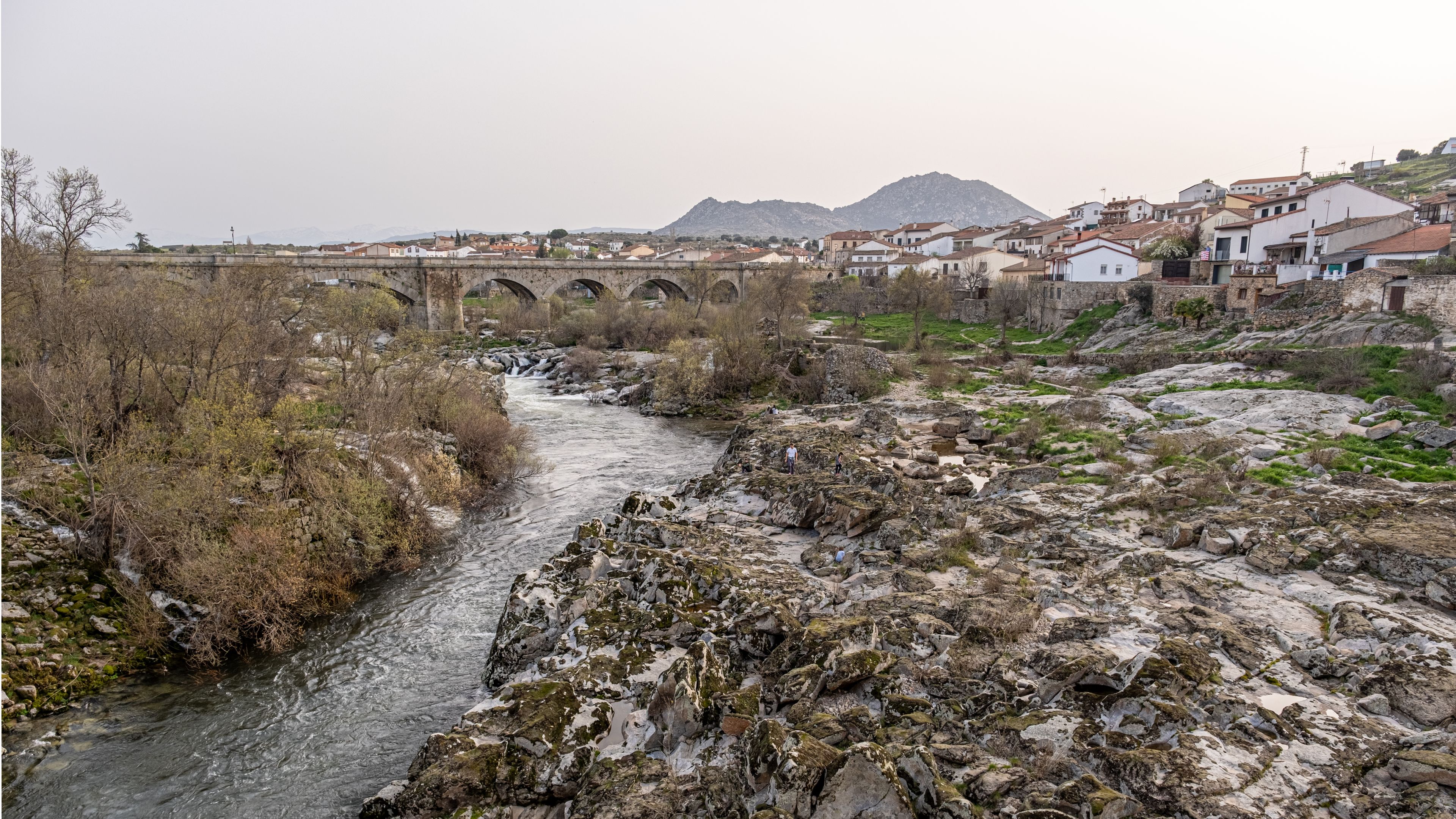 A river flowing under a stone bridge with a mountainous backdrop and a village with white houses.