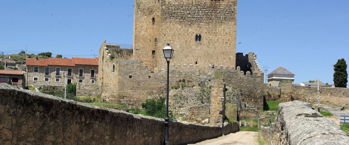 A historic stone tower and wall, a dirt path, and modern buildings in the background.
