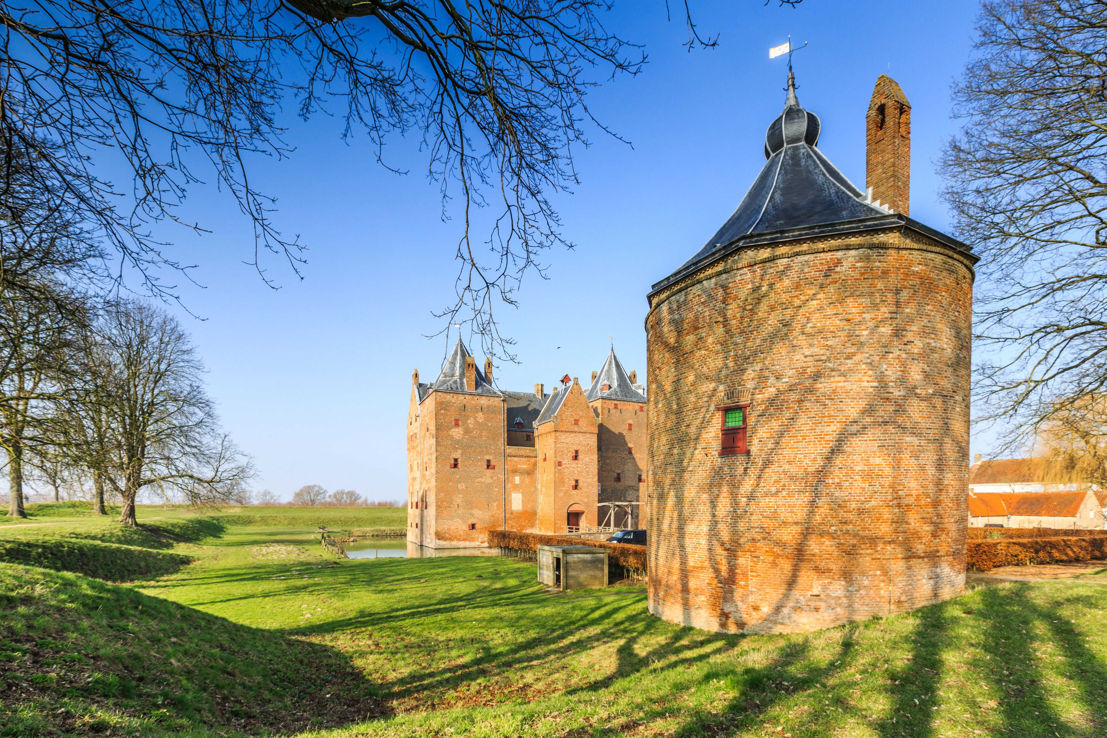 A historic brick tower with a conical roof, a large castle in the background, and a grassy field with a small pond.