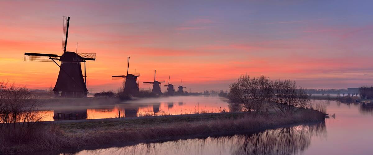 Windmills silhouetted against a sunset, a calm river, and a misty landscape.