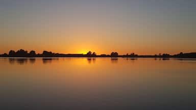 Um lago calmo ao pôr do sol, com céu limpo e silhuetas de árvores e uma linha de costa distante.
